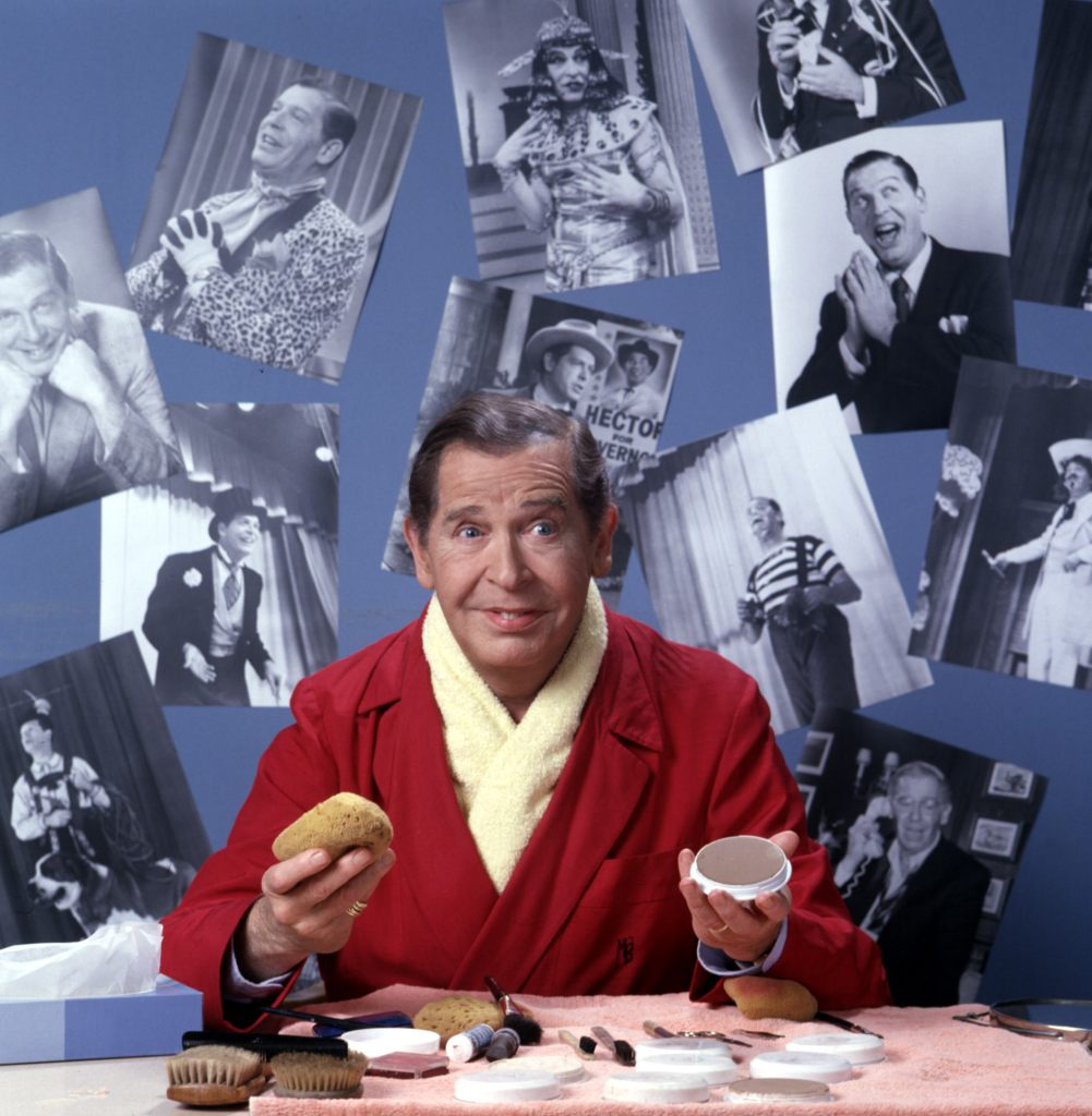 Milton Berle at the makeup table in front of pictures of himself in character in the 1970s