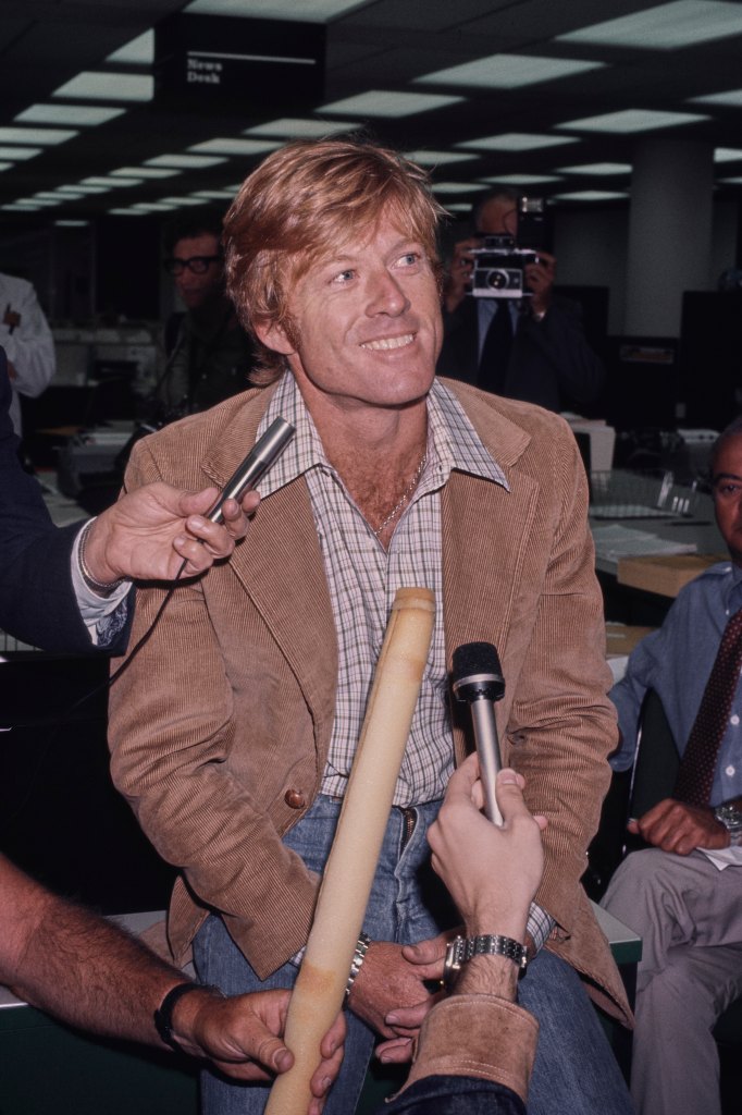 American actor Robert Redford, wearing a brown corduroy blazer over a checked shirt, is surrounded by microphones and what appears to be a baguette as he is interviewed in a recreation of The Washington Post's offices, on the set of 'All the President's Men,' filmed at Burbank Studios in Burbank, California, 1976. The political thriller based on the Watergate scandal, directed by Alan J Pakula, starred Redford as Bob Woodward