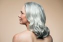 Mature woman with medium length, silvery, grey hair with her naked back and shoulders turned towards camera in front of a beige background looking to her left, profile.