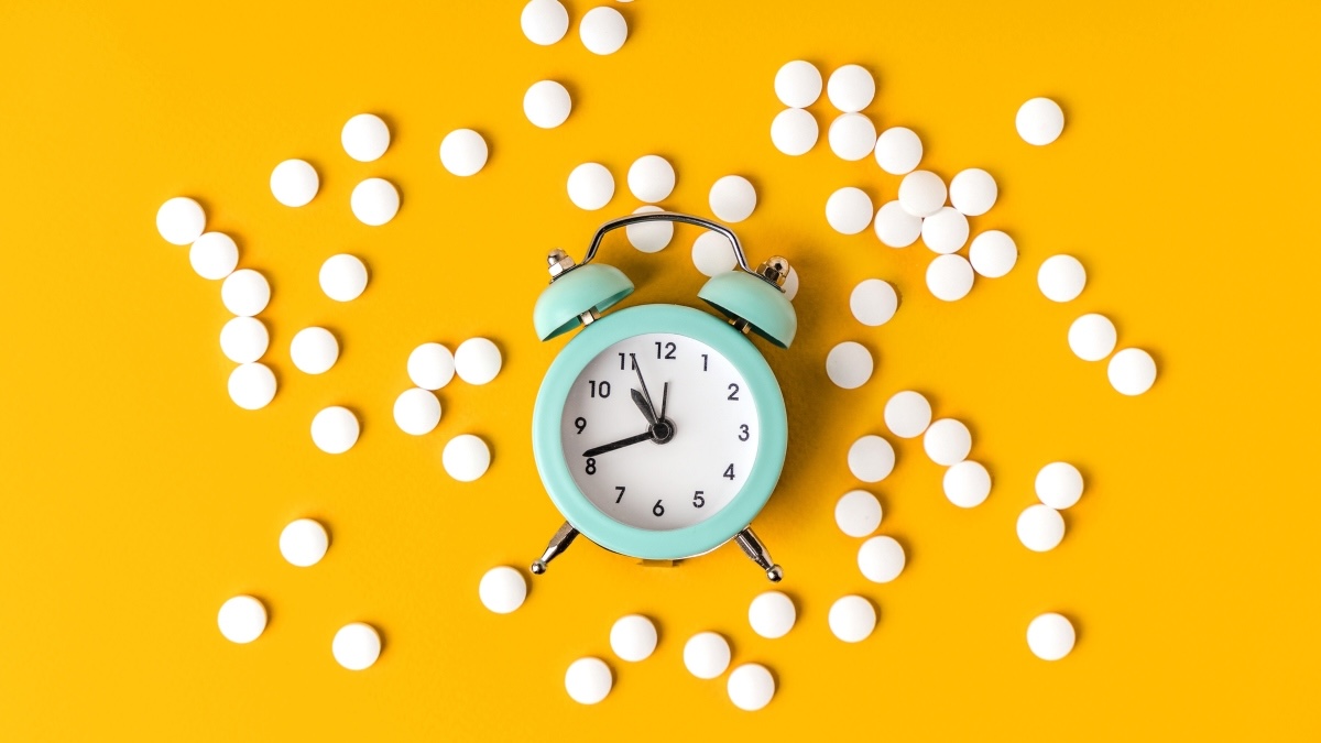 A clock, representing the best time to take vitamins, surrounded by white pills against a yellow background