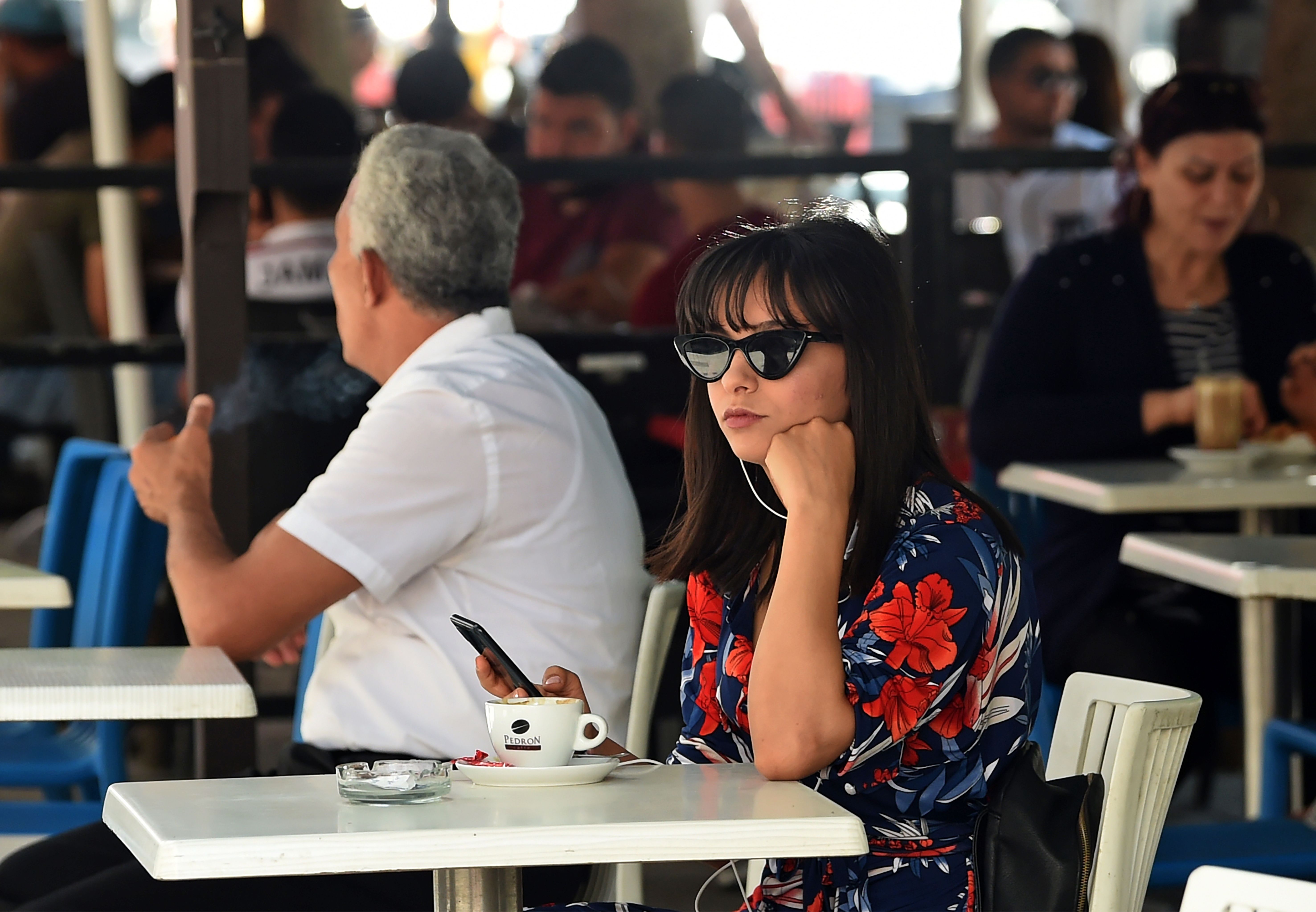 A woman sits at a cafe with her headphones in, wearing sunglasses.