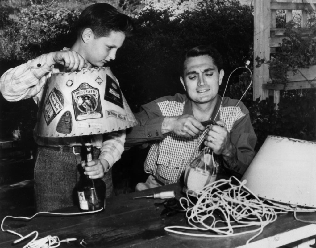 1945: Actor Robert Alda (1914-1986) shows his son Alan how to fashion homemade lamps from wine and liquor bottles outside their home in California. 