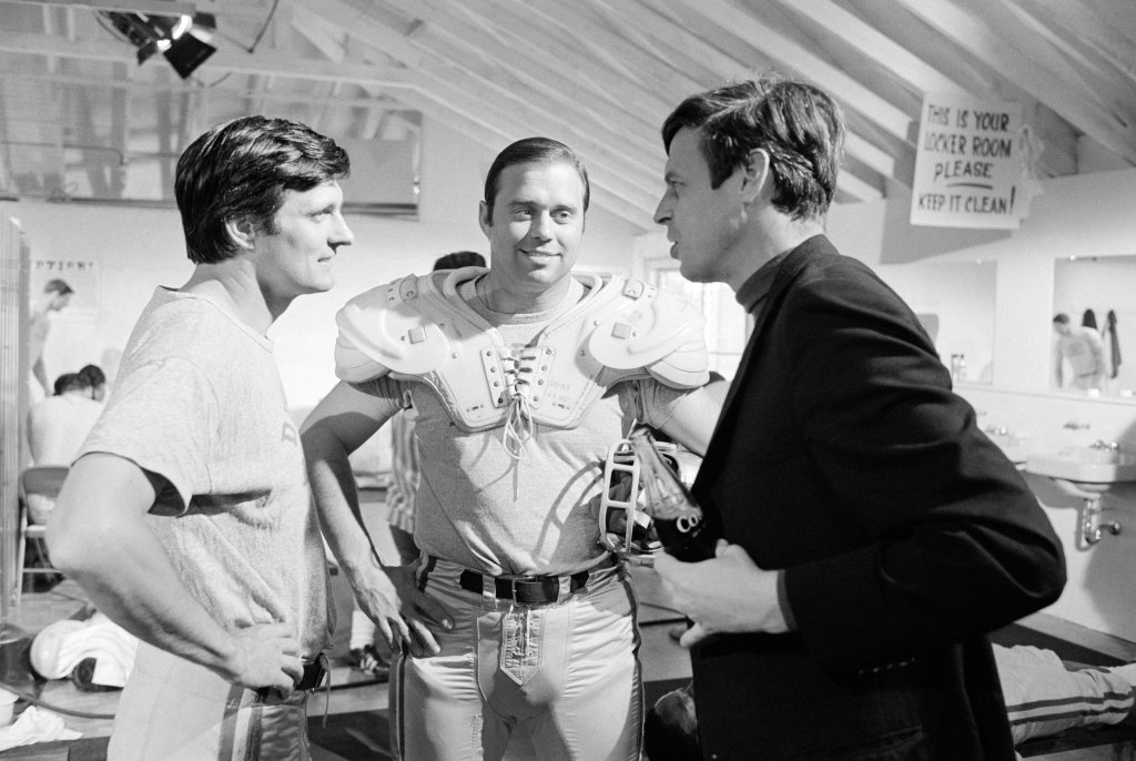 Author George Plimpton confers with Alan Alda on the set of the 1968 comedy Paper Lion, a film adaptation of Plimpton's best-selling book. Alda plays Plimpton's screen counterpart in a story of the author's masquerade as a professional quarterback with the Detroit Lions. Various NFL coaches and players, including Lions' offensive guard John Gordy (c), will also appear in the film.