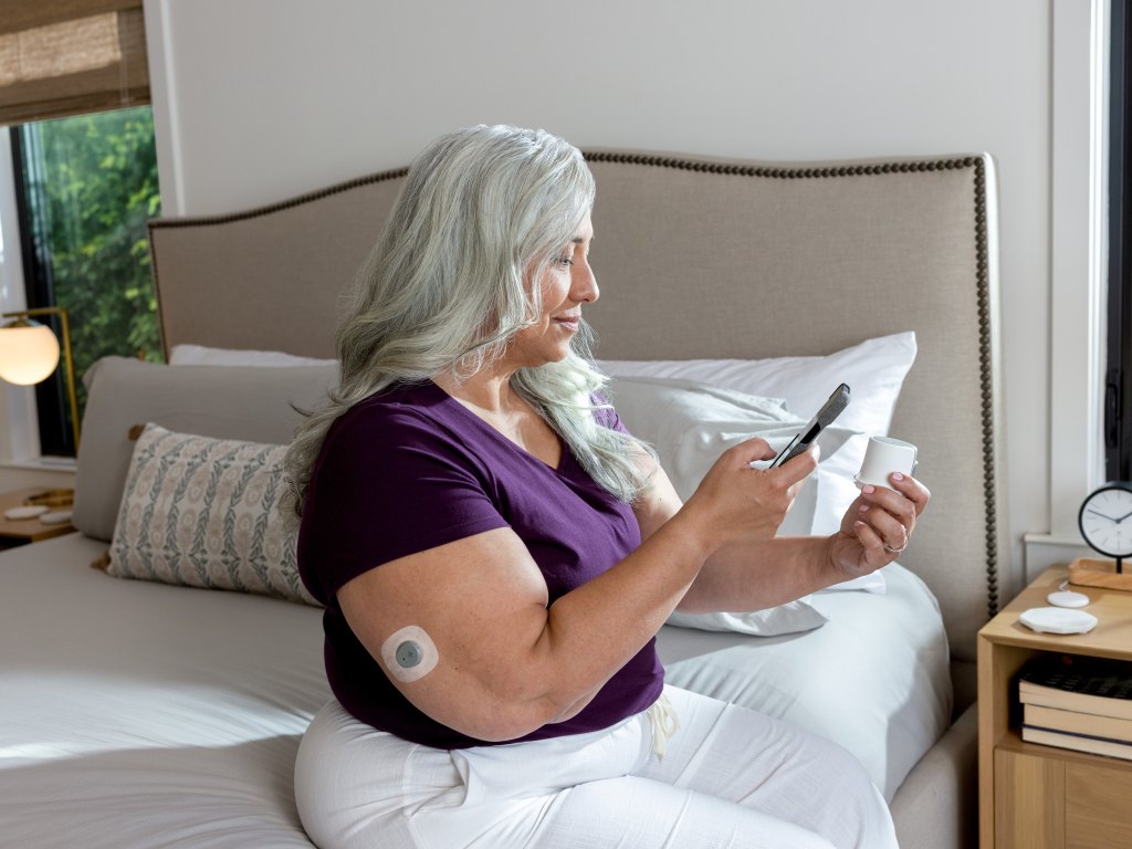 A woman sits on her bed and reads her phone to learn her blood-sugar level from wearing a Dexcom continuous glucose monitor on her arm.