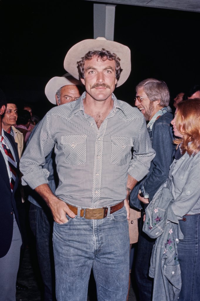 American actor Tom Selleck, wearing an open collar checked shirt, jeans and a cowboy hat, among guests attending an event, United States, circa 1980. 