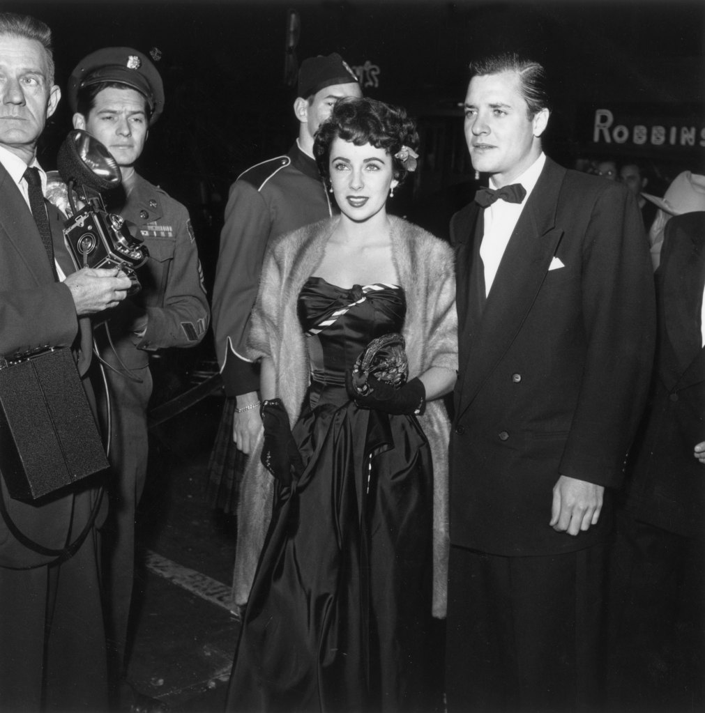 Elizabeth Taylor, wearing an evening gown with a fur coat, arrives at a film premiere with American actor Richard Long (1927 - 1974), wearing a tuxedo