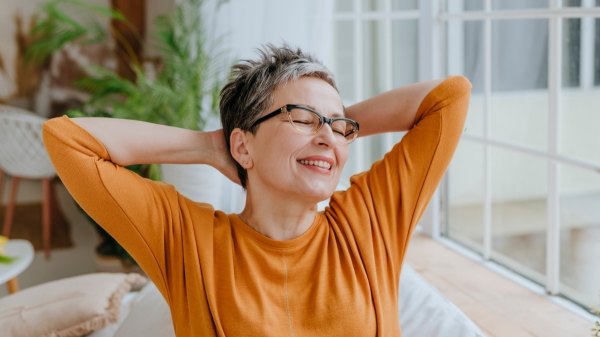A woman in an orange shirt with her hands behind her head smiling after doing a vagus nerve reset