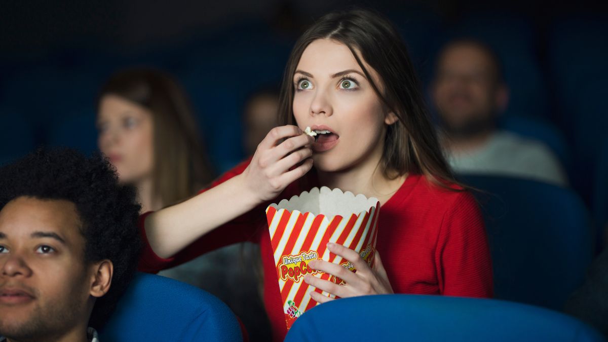 Woman sits in a movie theater eating popcorn, watching a movie to get scared