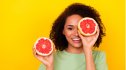 Woman holding a sliced citrus fruit, which is good for eye health, in front of one eye against a yellow background
