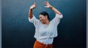 A midlife woman happily dances and stretches in front of a pretty blue wall to help prevent dementia