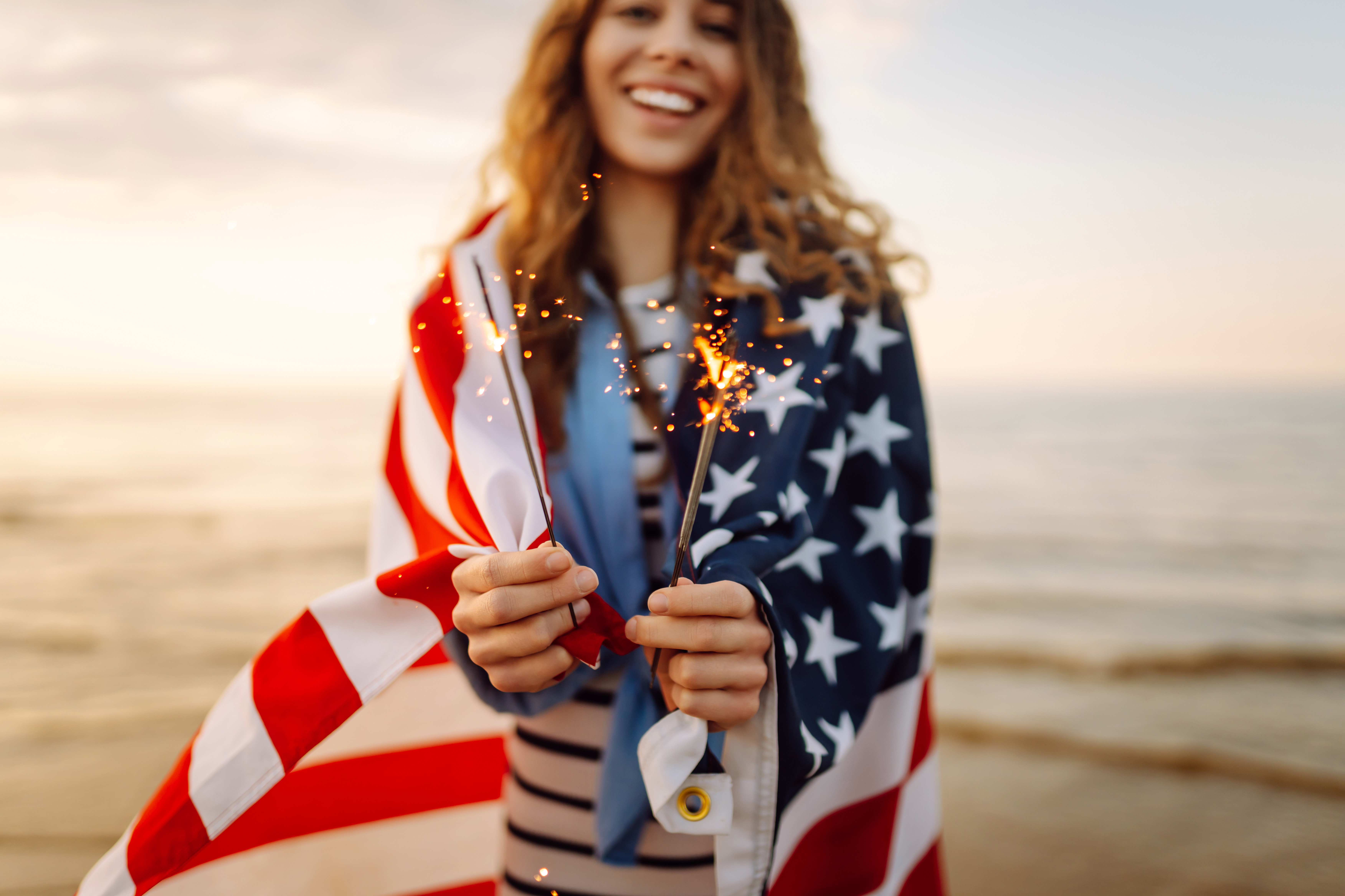 Young woman wrapped in American flag holding sparklers on sunset beach. Beautiful woman enjoying sunset. Concept of independence, celebration.