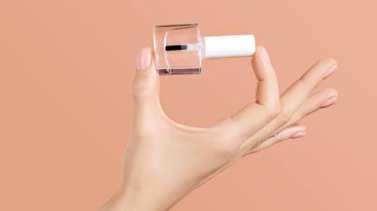 Uses for clear nail polish: Woman's hand holding a clear flask of nail polish. Applying the transparent varnish. Beige background, fingernail care at home concept. Image for nail salon or manicure artist promotion.